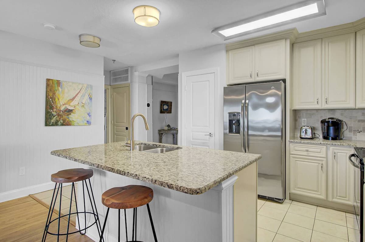 Kitchen from opposite angle showing bar stools, French-door fridge with ice maker, sailboat canvas art, and coffee station with Keurig