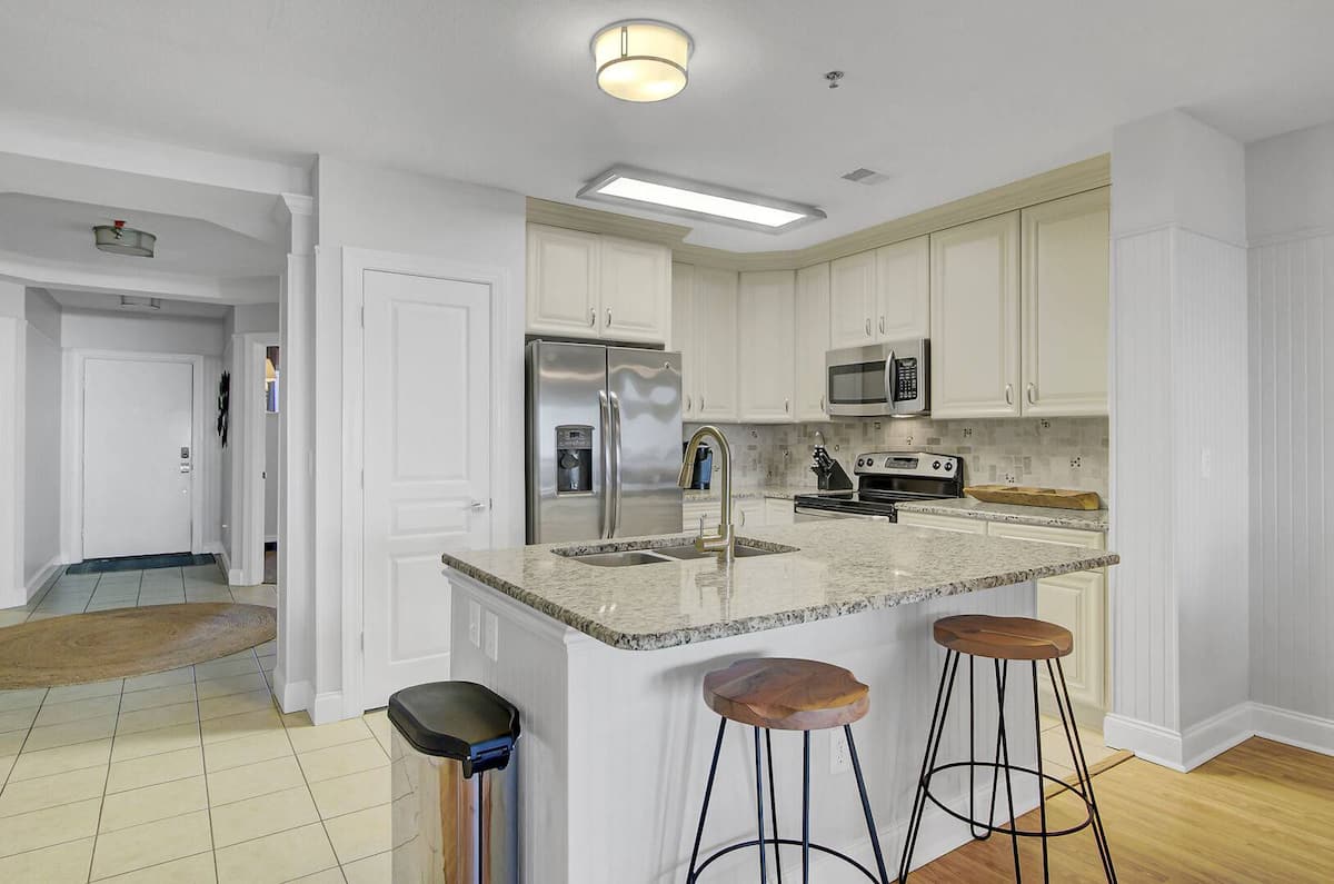 Kitchen island close-up with granite counters, industrial wood-and-metal bar stools, stainless French-door refrigerator, and hallway to bedrooms