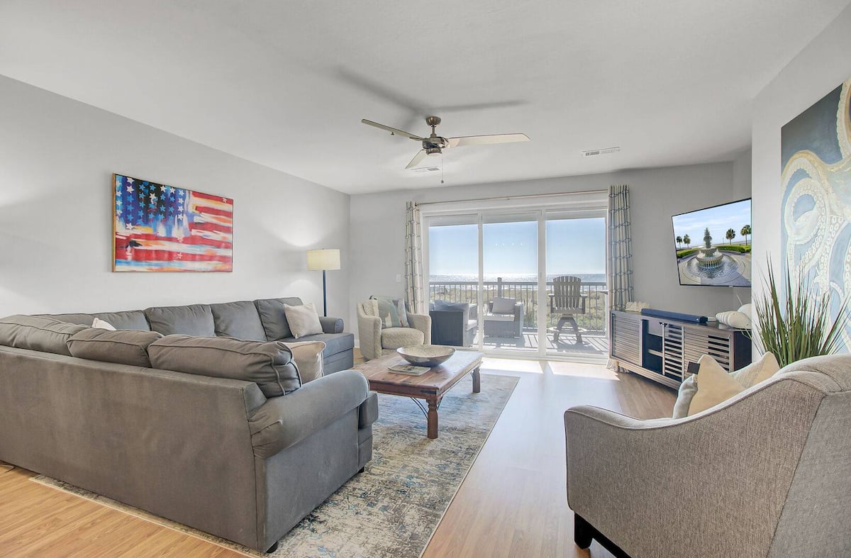 Living room with American flag canvas art, L-shaped gray sectional sofa, ceiling fan, and sliding doors open to oceanfront balcony with Adirondack chairs