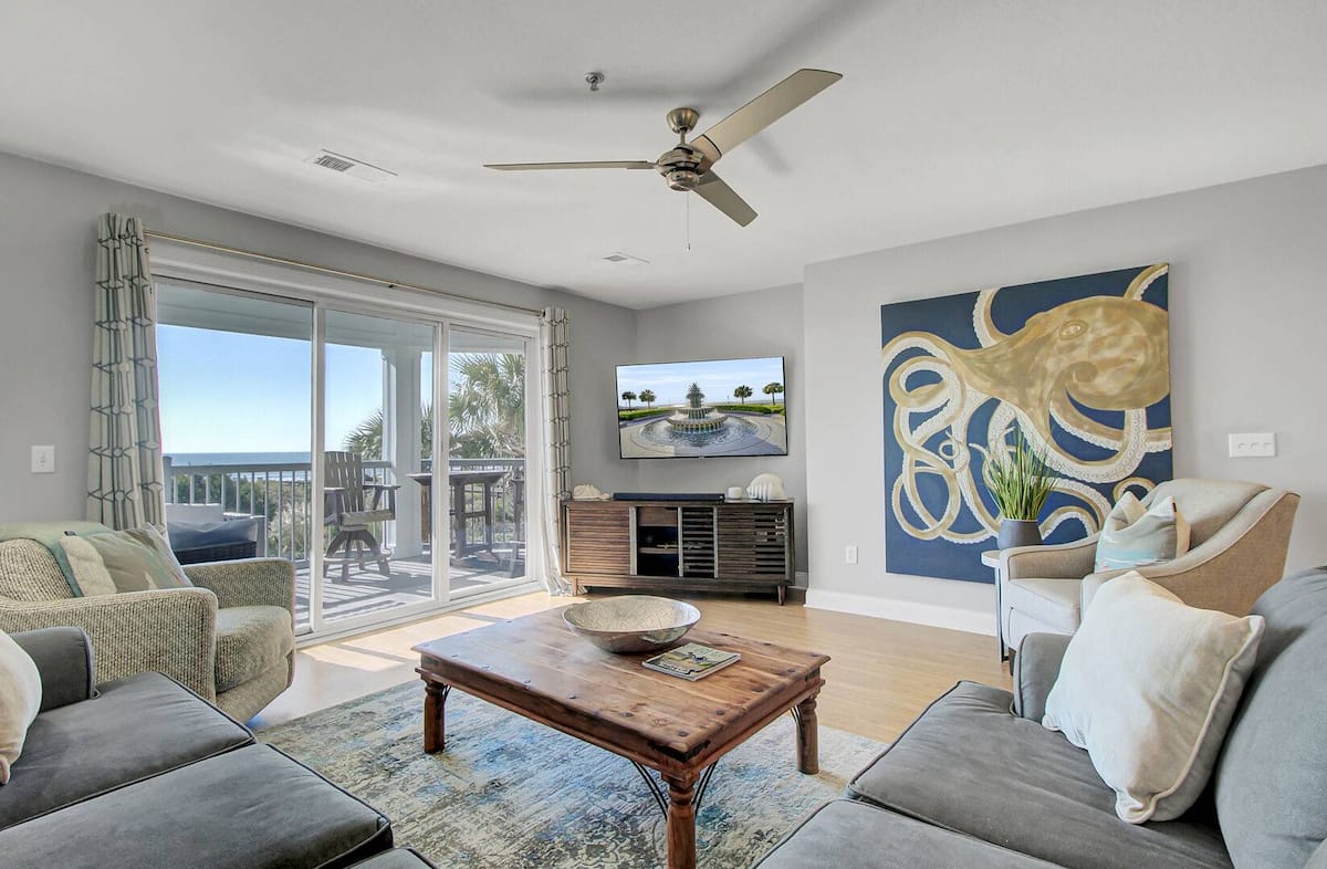 Living room with coastal decor, large golden octopus painting, ceiling fan, rustic coffee table, and ocean view through sliding glass doors to balcony