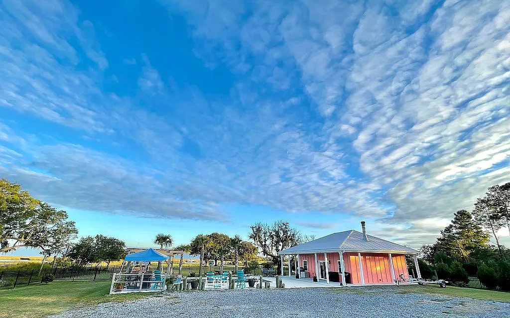 The full Bulls Bay compound at blue hour — salmon-pink playhouse with turquoise Adirondack chairs, fire pit, live oaks, and dramatic Lowcountry sky